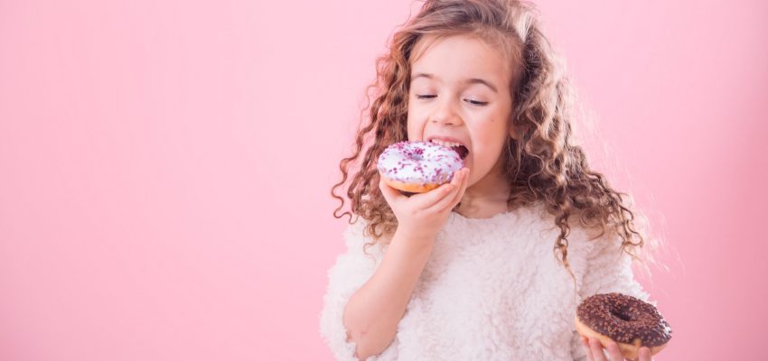 Portrait of a little joyful cute curly girl who eats donuts, on a pink background, a place for text