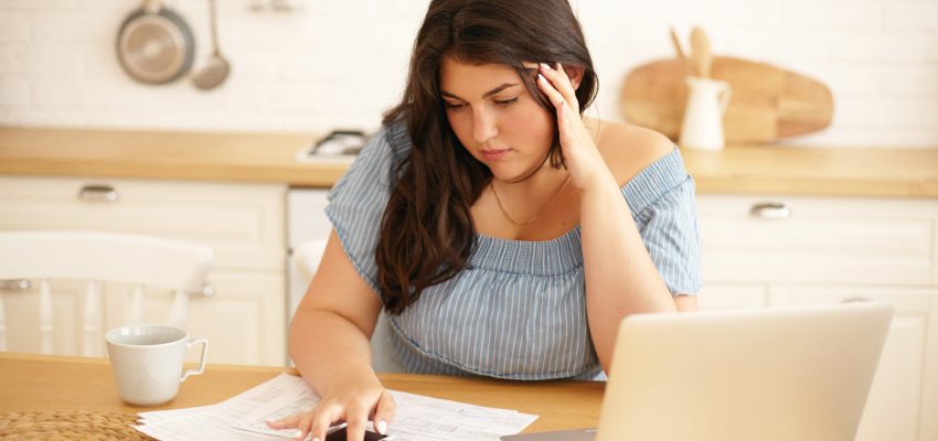 Concentrated young plus size woman sitting at table with papers and portable computer, using calculator, having focused serious facial expression, touching head, doing finances in stylish kitchen