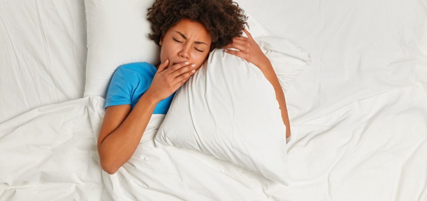 Horizontal shot of fatigue woman has not enough sleep, covers mouth while yawns, lies on white pillow under blanket, has sleepy look, difficult to wake up very early. Female has sleeping disorder